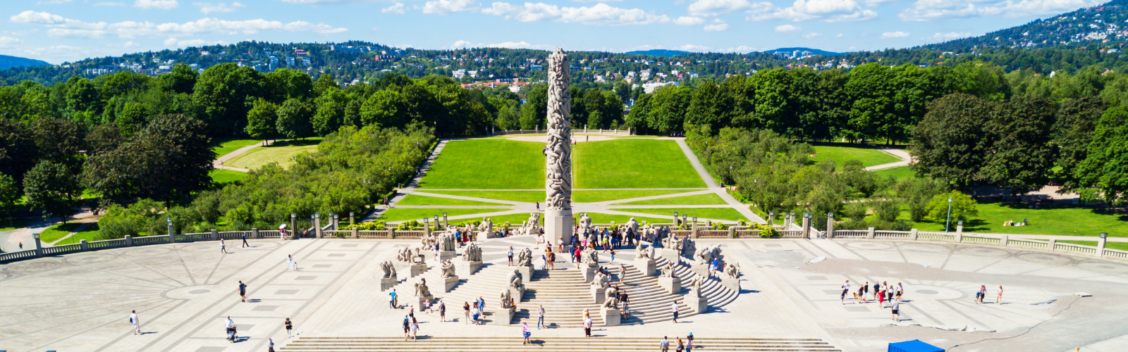 A tall, cylindrical stone sculpture stands in front of a green lawn, with many hills in the distance. 